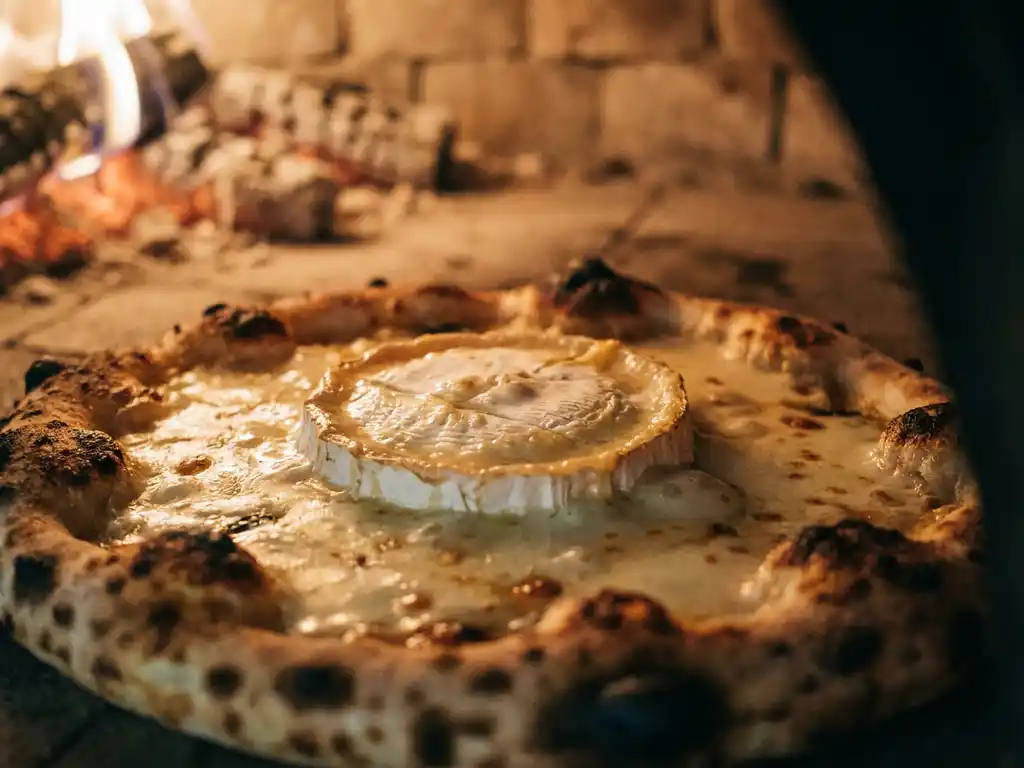 Smeltende geitenkaas op een houtgestookte pizza met goudbruine korst en zichtbare steenoven op de achtergrond.